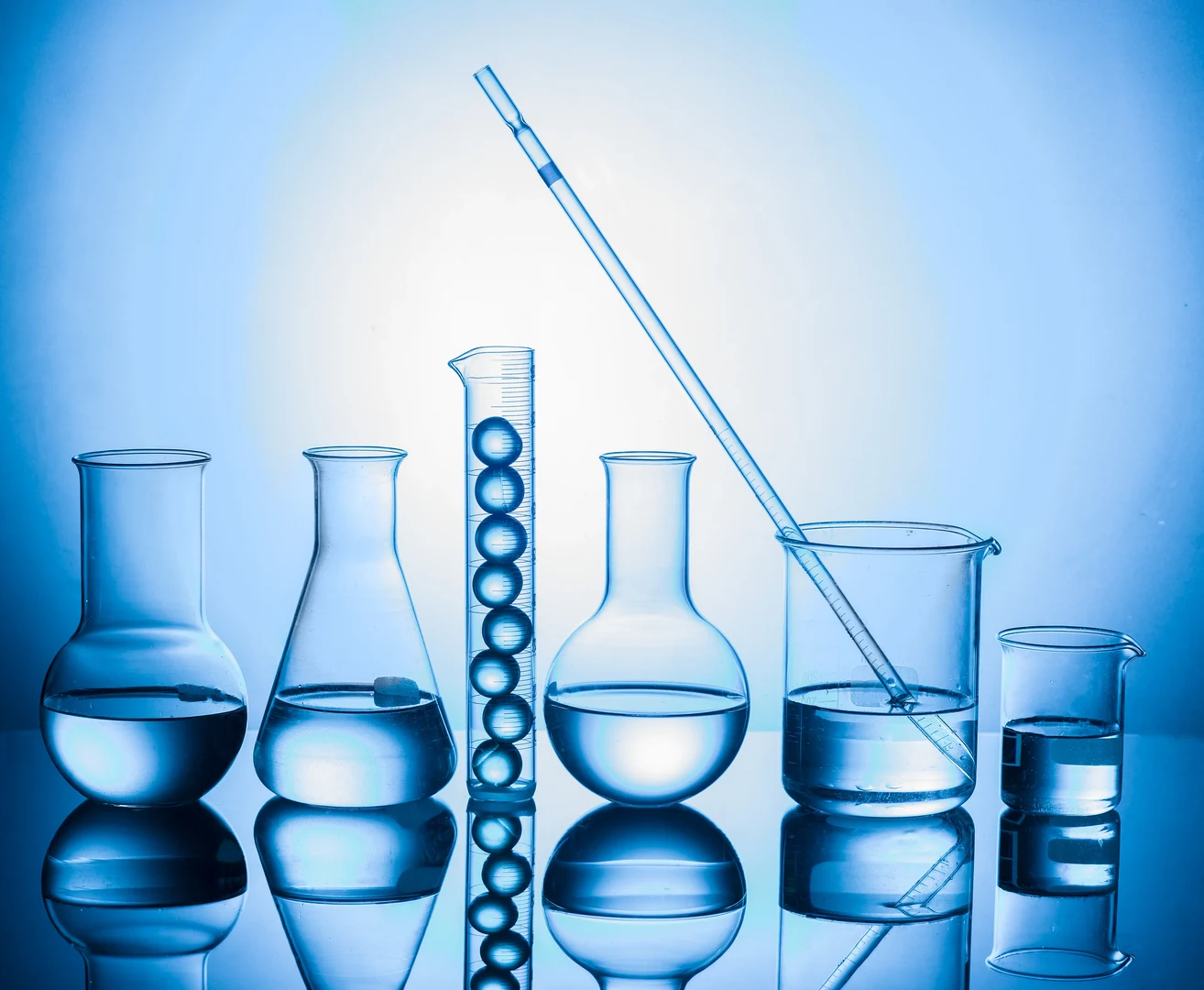 Eine Wissenschaftlerin und ein Wissenschaftler arbeiten hinter einer Glasfassade und mischen Chemikalien mit Großgeräten.